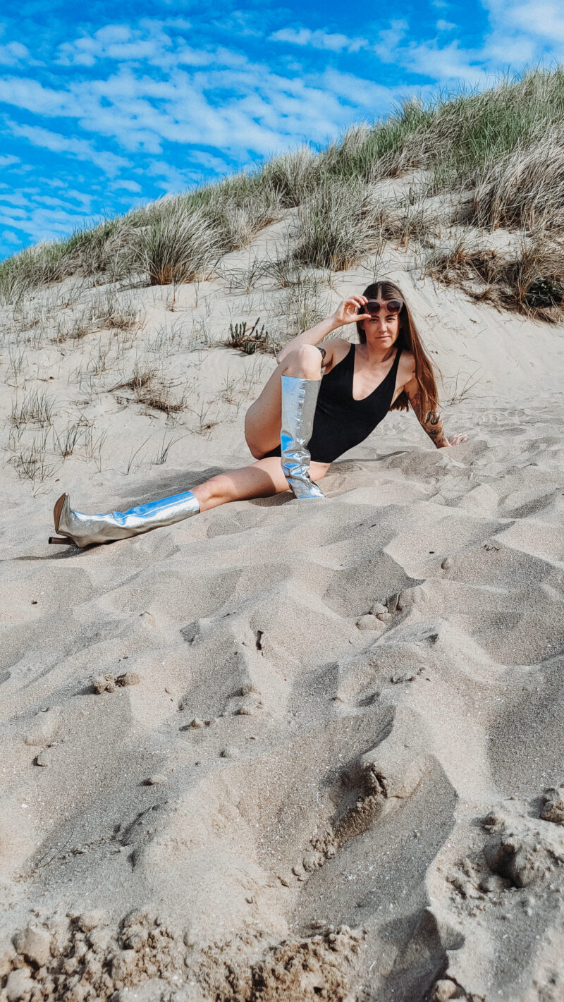 Woman sitting in the dunes peeking behind her sunglasess wearing black bodysuit Nanette Lindeman